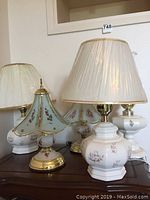 Four decorative table lamps arranged on wood furniture with wall behind. Three ceramic floral lamps with shades and one stained glass shade lamp. Plastic still on some shades.
