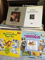 Four vintage vinyl albums including Louis Armstrong jazz, Winnie the Pooh and Tigger Too, The Aristocats, and others arranged on and in a shelf.