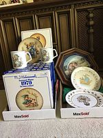Photo of 2 M.J. Hummel ceramic mugs and 2 annual plates on display, mugs sitting on one plate box and plates visible both inside and outside boxes.