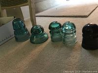 Five green glass insulators of various designs and a darker colored insulator on carpet.