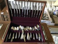 Photo showing the entire flatware set in the wooden case with burgundy velvet lining, including knives, forks, spoons, and specialty utensils arranged in compartments.