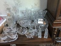 Full view of 11 pieces of cut glassware including decanters, salt and pepper shakers, bowls and vases arranged on wooden surface.