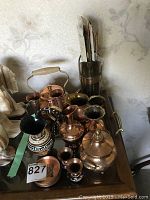 Overview of assorted copper and metal pitchers, tea pot, vessels, and coasters arranged on a tray on a wooden table.
