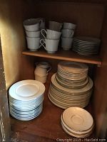 White porcelain cups, stacked saucers, luncheon plates, creamer and sugar bowl on wooden shelves.