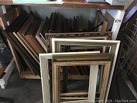 Multiple assorted picture frames stacked and leaning against shelving and walls in storage area. Pictures show frames of various sizes, shapes, and styles with wood, painted, and decorative trims.