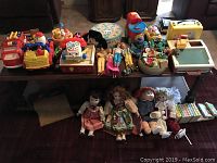 Wide view of all toys and dolls arranged on table and floor including plastic vehicles, dolls, books, and boxes.