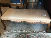 Upscale photo of upholstered wooden ottoman in beige fabric with subtle pattern and nailhead tacking, curved legs and scalloped front edge