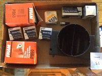 Top-down view of various automotive parts including two FRAM PB50 oil filter boxes, Cub Genuine Parts boxes, a NAPA filter box, a black automotive housing part, and switches arranged in a cardboard box.