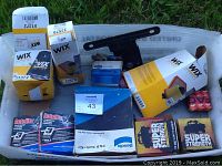 View of automotive parts including WIX oil and air filters, Autolite platinum spark plugs, carburetor kit, Super Strength boxes, and a black metal bracket in white container on grass.