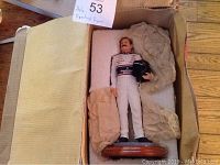 Dale Earnhardt #3 NASCAR collectible figure in white racing suit holding black helmet, placed in cardboard box with packing paper.