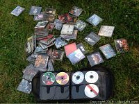 Photo showing a pile of empty CD jewel cases and some loose CDs organized in a black booklet, placed on grass.