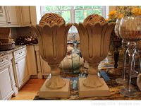 Two cream-colored resin planters with floral petal design on square bases, standing on kitchen counter near window with other decor