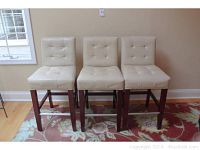 Front view of three off-white vinyl upholstered bar stools with dark wood legs and button tufted backs and seats, placed on a floral patterned carpet.