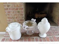 Three items displayed on brick hearth: two white ceramic urns with lids and decorative handles, and a beige resin planter with pedestal and decorative features