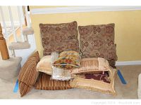 Photo of a pile of 9 decorative throw pillows in various patterns and sizes on carpeted floor near stairs and wall.