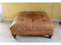 Front view of a large brown leather ottoman with tufted top and pin head accents on base, showing some fading and wear.