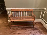 Wooden hall bench with spindled backrest and armrests, wicker seat, standing on hardwood floor against wainscoted wall.
