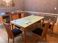 Dining table with extension leaf surrounded by six wooden chairs with black cushions, hardwood flooring and gray walls in background.