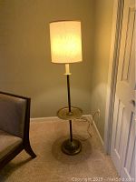 Floor lamp illuminated showing fabric drum lampshade and integrated brass-colored table shelf on metal stand with decorative base near sofa.