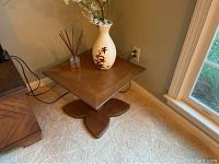 End table placed against a wall beside a window on carpet. Table has a brown wood finish with a floral vase and reed diffuser on top.