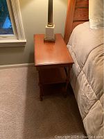 Photo of a wooden end table placed beside a bed with a lamp on top, showing the top surface and two lower shelves.