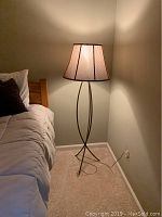 Floor lamp standing next to a bed with an off-white fabric shade and metal frame with double-loop base design.
