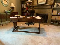Front angled view of walnut desk with papers on top in a room with tiled floor, bookshelf and fireplace in background