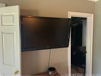 Sharp television mounted on wall showing front view with screen off, environment includes beige wall and a partially open door.