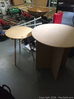 Shows both round pressed wood tables together in a garage setting, giving a general scale and height comparison between the two.