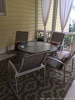 Four matching beige metal frame outdoor chairs with patterned mesh fabric and red flower decorative pillows arranged around a round glass table on a patterned rug on screened porch