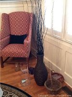 Large dark brown wicker vase with tall sticks by a patterned armchair and other glass vases on wood floor