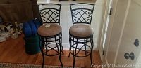Two swivel bar stools with metal frames and brown suede seats shown against a wall with hardwood flooring. Both stools have curved and decorative metal backrests and round seats.