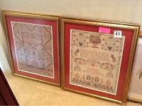 Two framed print cross stitch samplers leaning against the wall, showing gold frames and red mats.