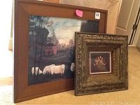 Two framed paintings displayed leaning on carpet: larger folk art landscape with sheep in wooden frame and smaller floral painting in ornate carved frame.