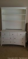 Photo of white wooden dresser with seven drawers and silver knobs, bookshelf hutch on top with two shelves.