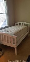 White twin bed frame with pillowtop mattress against wall near window, showing side and footboard.