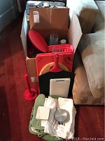 Photo showing cardboard box filled with Pyrex glass bowls with red lids, cutting boards leaning outside the box, napkins, oven mitt, coasters, and paper towel holder.