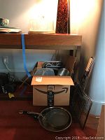 View of cardboard box filled with various baking containers with lids, grill racks leaning against wall, two frying pans on floor in front.