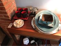 Table with four black and red inside ceramic cups on a red cloth, small stack of Christmas themed saucers, glass candy jar, and Pier 1 Hibiscus serving bowl and platters with a large stainless steel platter underneath.