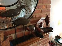 Six carved bird decoys arranged on a wooden shelf in front of a brick wall and mirror, showing both the three red shorebirds and three larger pedestal birds.