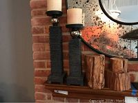 Pair of tall iron candle holders with silver spherical accents and cream candles on a mantle in front of a mirror with red brick background.