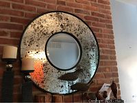 Front and angled views of the round antique finish mirror showing its distressed reflective surface and black outer frame against a brick wall background.