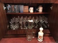 Glassware collection inside a cabinet including beer pint glasses, wine glasses, champagne flutes, coffee mugs, a clear glass pitcher, and a ceramic whiskey decanter.