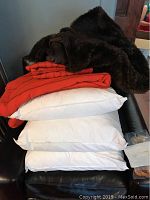 Stack of three white square pillows under orange wool blanket and dark brown faux bear fur blanket, placed on a black chair.