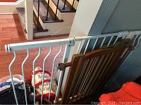 Photo showing two pet gates: a white metal gate and a brown wooden adjustable gate leaning against a staircase railing.