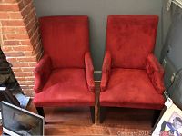 Two red suede dining arm chairs side by side with wood floor and brick wall background, showing overall shape and fabric color