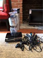 Sony PlayStation 3 console with controllers, wired microphones, and stacks of physical game cases on floor near TV.