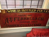 Two wooden signs displayed on a window sill: top curved wooden wine sign painted in yellow text and bottom rectangular painted farming company sign in red with black stencil text.