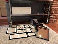 Photo showing multiple picture frames arranged on carpet under a metal shelf, including black and brown framed pieces and unopened box sets.