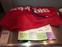 Red Washington Capitals hockey jersey lying flat showing several black ink player signatures and a pamphlet indicating autograph areas and signatories.